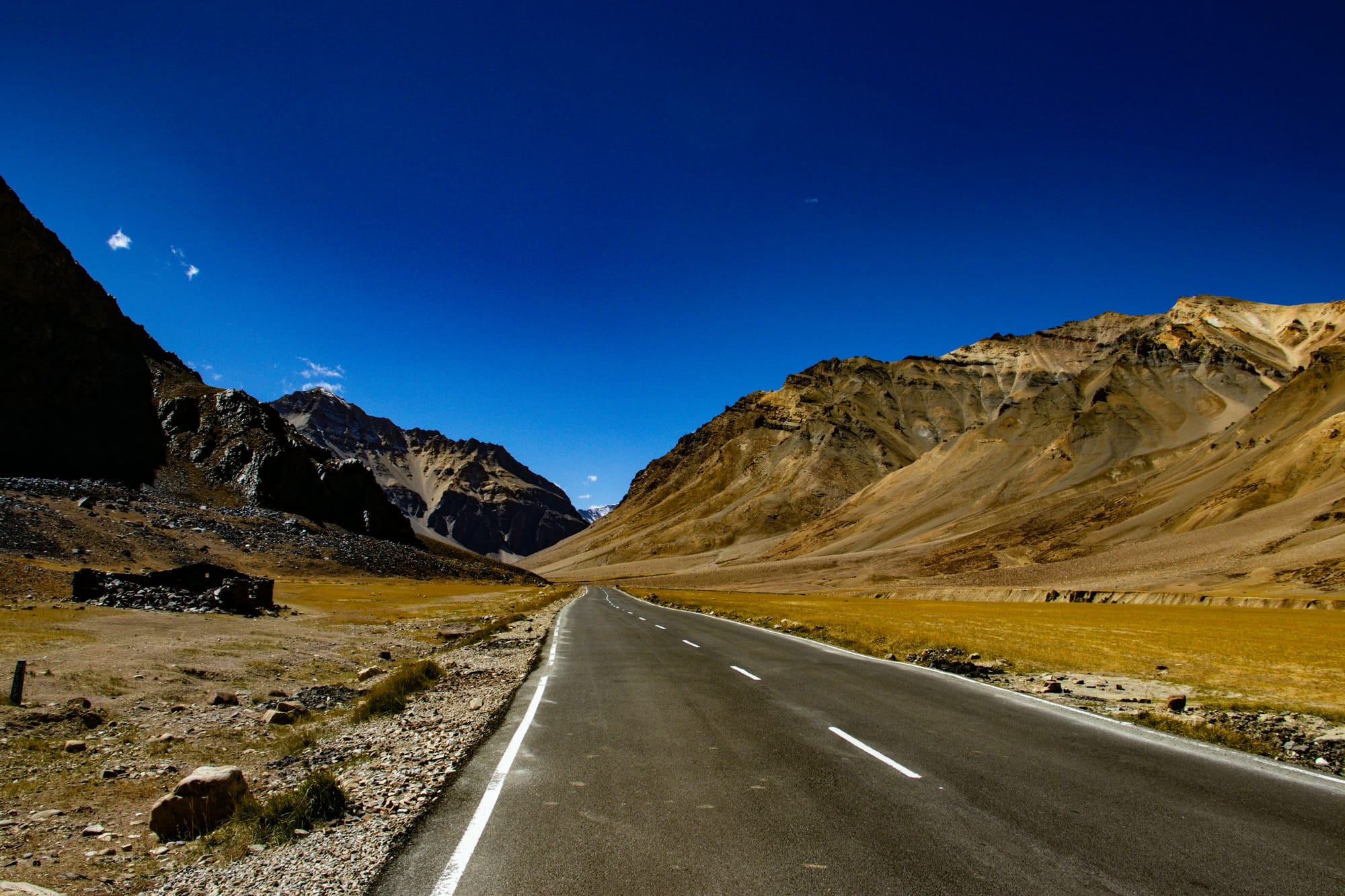 An empty highway running through the bare golden mountains of Ladakh under a deep blue sky